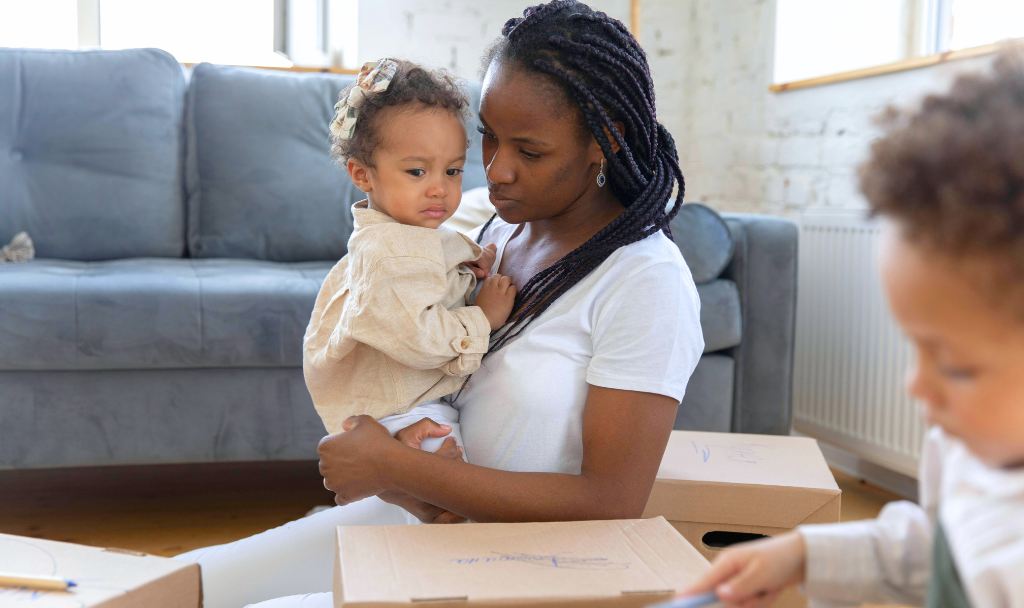 Mommy Burnout. Image of a mother holding a crying child as another sits in the foreground, a couch and windows in the background.